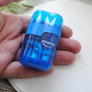 May include: A blue Maped pencil sharpener with a transparent body. The sharpener is held in a hand and has the brand name printed on the side. The sharpener is made of plastic and has a rectangular shape.