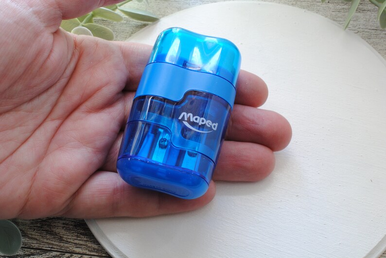 May include: A blue Maped pencil sharpener with a transparent body. The sharpener is held in a hand and has the brand name printed on the side. The sharpener is made of plastic and has a rectangular shape.