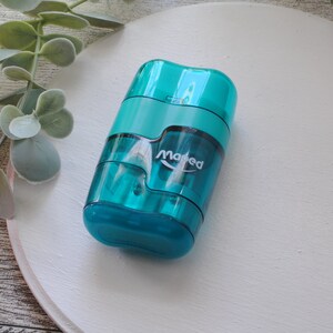 May include: A teal-colored pencil sharpener with the brand name "Maped" printed on the side. The sharpener has a transparent body, allowing visibility of the pencil shavings. The design is compact and features a dual-hole system for different pencil sizes.