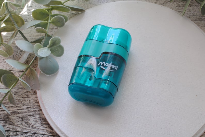 May include: A teal-colored pencil sharpener with the brand name "Maped" printed on the side. The sharpener has a transparent body, allowing visibility of the pencil shavings. The design is compact and features a dual-hole system for different pencil sizes.
