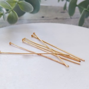 May include: A group of gold-colored head pins, also known as jewelry making pins, are arranged on a white surface. The pins are straight and have a rounded head.