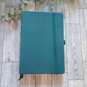 May include: A teal-colored notebook with a textured cover, elastic closure, and ribbon bookmark. The notebook is rectangular and closed, with a matching teal elastic band running vertically. The background is a light-colored wooden surface with a sprig of greenery.