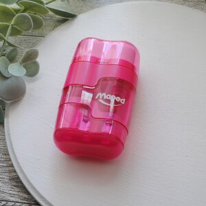 May include: A bright pink Maped pencil sharpener with a clear plastic body. The sharpener has a rectangular shape with rounded edges and a small white logo. The sharpener is on a white surface with green foliage in the background.