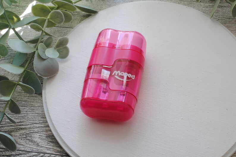 May include: A bright pink Maped pencil sharpener with a clear plastic body. The sharpener has a rectangular shape with rounded edges and a small white logo. The sharpener is on a white surface with green foliage in the background.