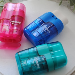May include: Three Maped pencil sharpeners in pink, blue, and teal. The pink sharpener has a removable tray. The blue sharpener has a white eraser. Each sharpener has the Maped logo printed on the side. The sharpeners are on a white surface.