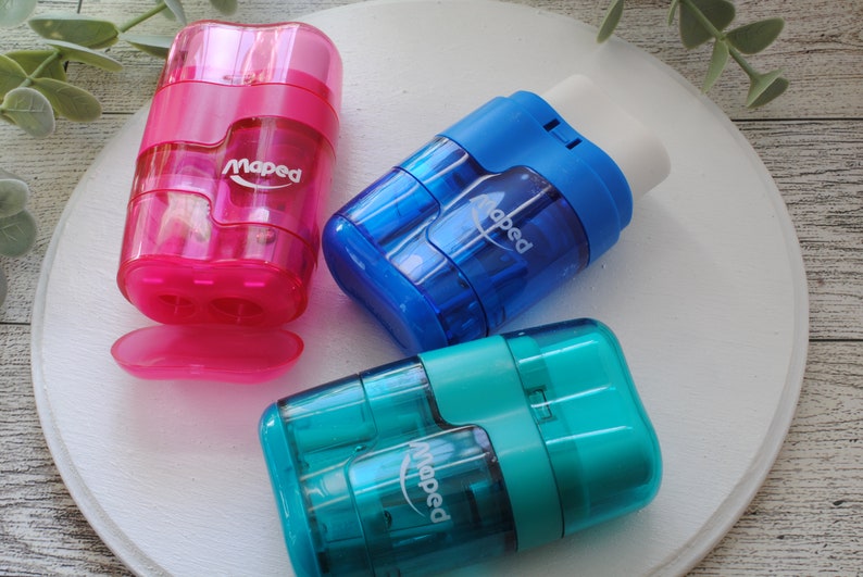 May include: Three Maped pencil sharpeners in pink, blue, and teal. The pink sharpener has a removable tray. The blue sharpener has a white eraser. Each sharpener has the Maped logo printed on the side. The sharpeners are on a white surface.