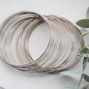 May include: A collection of silver-colored bangles, stacked and intertwined, creating a layered effect. The thin, circular bracelets are arranged on a white surface, with a sprig of greenery in the background.