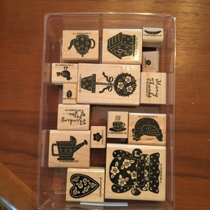 Könnte beinhalten: Ein Set mit 12 Holzstempeln mit verschiedenen Designs, darunter eine Teekanne, ein Vogel, ein Haus, eine Pflanze, eine Gießkanne, ein Herz, eine Tasse Tee und eine Frau mit einem Blumenmuster. Die Stempel befinden sich in einem durchsichtigen Plastikbehälter und sind mit dem Firmennamen "Stampin' Up!" beschriftet.