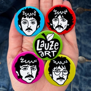 Könnte beinhalten: Fünf runde Ansteckbuttons mit schwarz-weißen Illustrationen von vier verschiedenen Gesichtern mit Schnurrbart und einer zentralen grünen Apfelgrafik mit dem weißen Text Lauze ART. Die Buttons sind in leuchtendem Blau, Rot, Pink und Gelb gehalten.