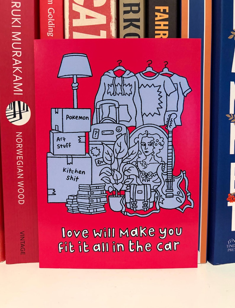 May include: A red greeting card featuring a light blue illustration of moving boxes, clothing, a lamp, a guitar, and a woman's portrait. The text "love will make you fit it all in the car" is at the bottom.
