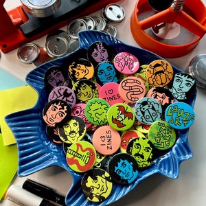 May include: A blue fish-shaped dish filled with colorful, round buttons featuring various illustrations and text. The buttons have images of faces, abstract designs, and the text "I <3 ZINES" and "Lauze ART". Button-making tools and markers are also visible.