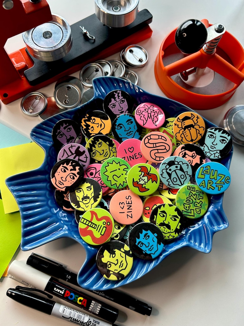 May include: A blue fish-shaped dish filled with colorful, round buttons featuring various illustrations and text. The buttons have images of faces, abstract designs, and the text "I <3 ZINES" and "Lauze ART". Button-making tools and markers are also visible.