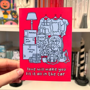 May include: A red greeting card featuring a black and white illustration of packed boxes, clothing, and a guitar. The text "love will make you fit it all in the car" is printed at the bottom.