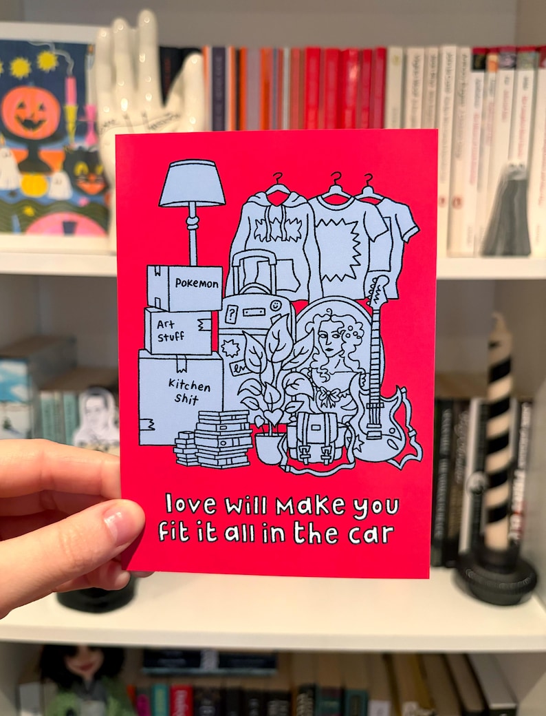 May include: A red greeting card featuring a black and white illustration of packed boxes, clothing, and a guitar. The text "love will make you fit it all in the car" is printed at the bottom.