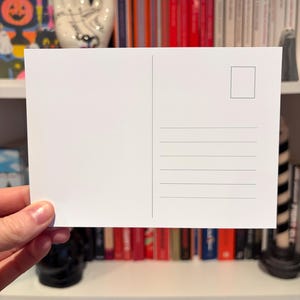 May include: A blank white postcard with a rectangular stamp box and horizontal lines for writing. The postcard is held in a hand, with a bookshelf filled with books in the background.