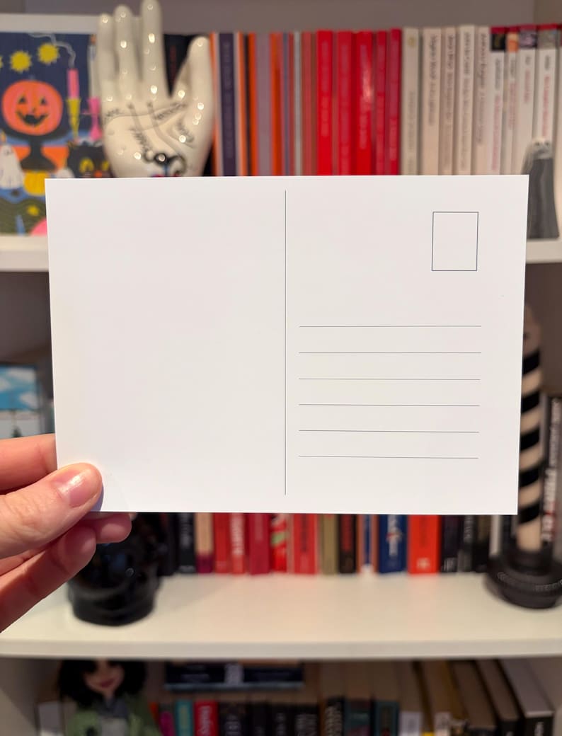 May include: A blank white postcard with a rectangular stamp box and horizontal lines for writing. The postcard is held in a hand, with a bookshelf filled with books in the background.
