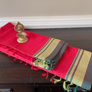 May include: A red cotton saree with a gold and green border. The saree is folded and displayed on a dark wooden surface. A small brass diya is placed on top of the saree.