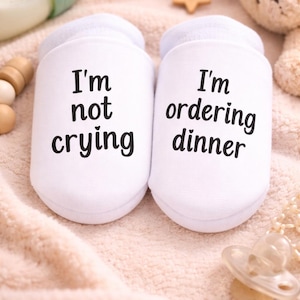 May include: White baby socks with the phrases "I'm not crying" and "I'm ordering dinner" printed in black. The socks are on a soft, peach-colored blanket, with a baby bottle, pacifier, and teddy bear in the background.