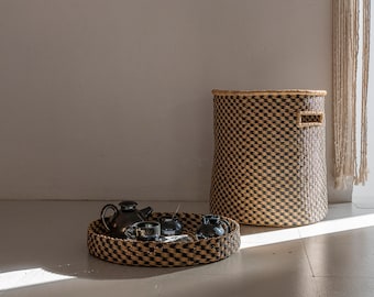 May include: A woven basket with a black and tan checkered pattern sits next to a woven tray with a black and tan checkered pattern. The tray holds a teapot and four small cups.