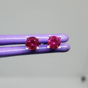May include: A pair of stud earrings featuring round, vibrant magenta gemstones set in silver prongs. The earrings are displayed against a soft, neutral background, highlighting their color and design. These earrings are a classic jewelry item.