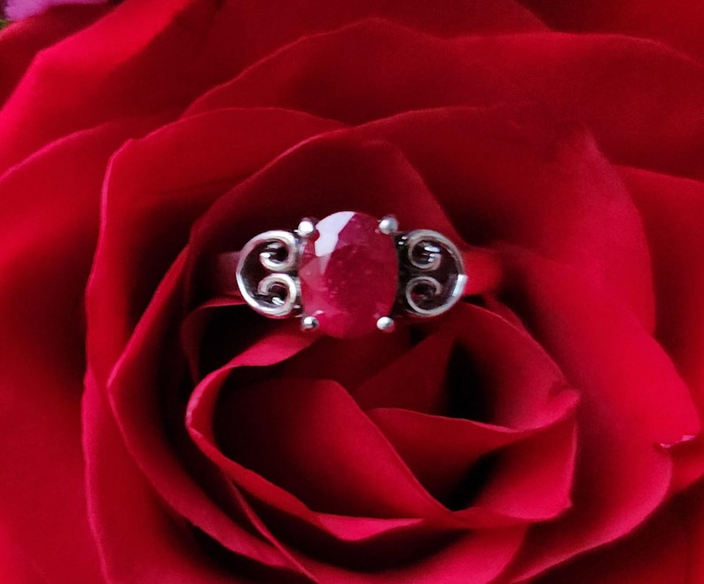 May include: A silver ring with a large, oval-cut ruby gemstone. The ring features decorative scrollwork on either side of the ruby. The ring is set against a backdrop of deep red rose petals, creating a striking contrast.