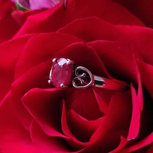 May include: A silver ring with a large oval ruby gemstone, set against the backdrop of a vibrant red rose. The ring features a delicate heart-shaped design on the side, adding a touch of elegance to the jewelry.