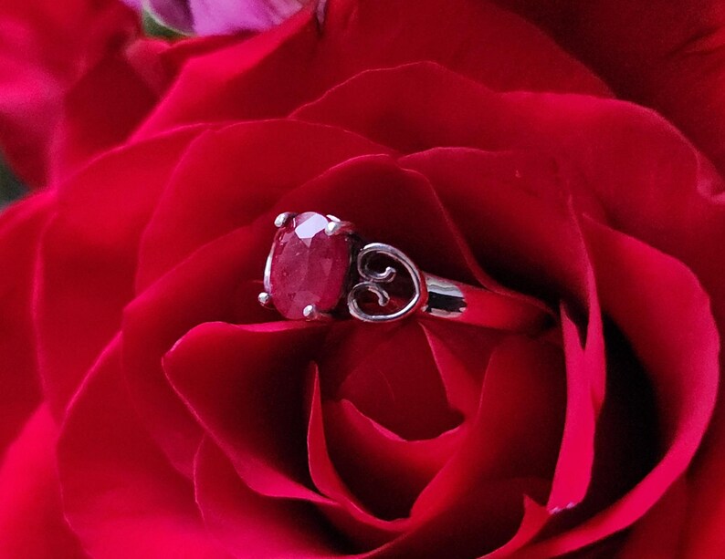 May include: A silver ring with a large oval ruby gemstone, set against the backdrop of a vibrant red rose. The ring features a delicate heart-shaped design on the side, adding a touch of elegance to the jewelry.