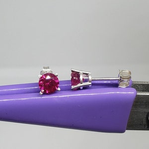 May include: Two silver stud earrings with vibrant pink gemstones. The gemstones are round-cut and set in a four-prong setting. The earrings are displayed on a purple surface against a light gray background.