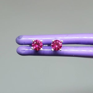 May include: A pair of stud earrings featuring round, vibrant pink gemstones set in a silver-colored metal. The earrings are held by a purple tool against a gray background, showcasing their color and design.