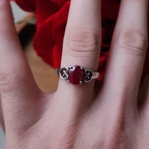 May include: A silver ring featuring an oval, deep red gemstone. The ring has a decorative filigree design on either side of the gemstone. The ring is shown on a finger, with a blurred red background.