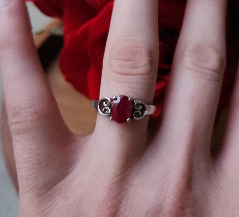 May include: A silver ring featuring an oval, deep red gemstone. The ring has a decorative filigree design on either side of the gemstone. The ring is shown on a finger, with a blurred red background.