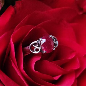 May include: A silver ring with a large, round, red gemstone, set against a backdrop of a deep red rose. The ring has an ornate, swirling design on the band.
