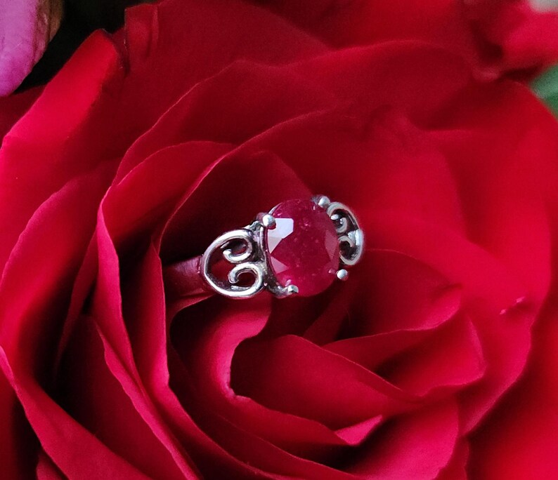 May include: A silver ring with a large, round, red gemstone, set against a backdrop of a deep red rose. The ring has an ornate, swirling design on the band.