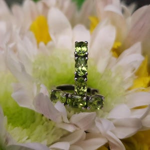 May include: A close-up of two rings with a floral design. The rings feature a row of oval-cut, light green gemstones. The rings are set against a backdrop of white and yellow flower petals, creating a soft, natural setting.