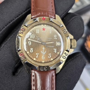 Puede incluir: Un reloj de pulsera vintage en tono dorado con una correa de cuero marrón. La esfera del reloj es de color marrón descolorido con un diseño de estrella roja y ancla. El reloj tiene una pantalla de fecha y un bisel giratorio.