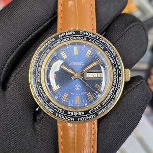May include: A vintage wristwatch with a blue face and gold-tone hands and markers. The watch has a brown leather strap and a bezel with city names. The watch face displays the day and date. The watch is held by a gloved hand.