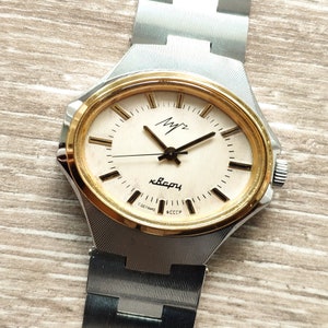 Puede incluir: Un reloj de pulsera plateado y dorado con una esfera blanca y agujas negras. La esfera del reloj tiene el texto "Мир" y "СССР" impreso en ella.