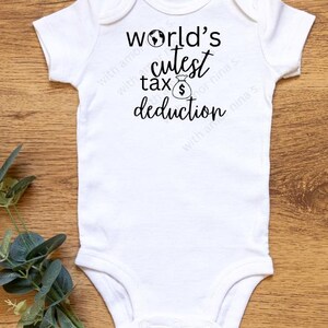 May include: A white baby onesie with short sleeves. The onesie has the text "world's cutest tax deduction" in black script. A small graphic of a globe and a money bag are included in the design. The onesie is on a wooden surface.