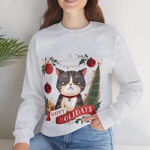 Op de afbeelding: Een grijs sweatshirt met een kerstthema met een chagrijnige kat met een rode halsband en een kleine kerstboom. De tekst "Happy Holidays" is gedrukt op een rode banner.