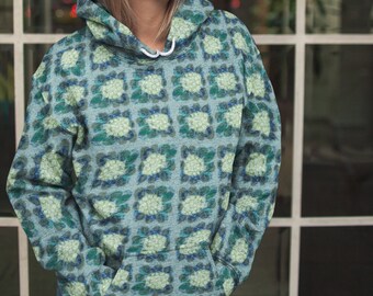 Könnte beinhalten: Hellblauer Kapuzenpullover mit einem sich wiederholenden Blumenmuster in Grün- und Weißtönen. Der Kapuzenpullover hat eine Kapuze mit Kordelzug und eine Vordertasche. Die Person trägt eine blaue Jeans.