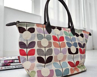 May include: A large tote bag with a colorful floral pattern in red, purple, blue, and orange. The bag has a cream background, brown leather handles, and a zipper closure. The bag is sitting on a white wooden floor.