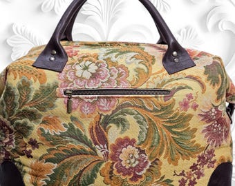 May include: A floral patterned handbag with a brown leather handle and accents. The bag features a zipper closure and a tapestry-like design with yellow, green, pink, and brown hues. The bag's dimensions are not visible.