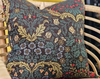 May include: Decorative throw pillow with a dark brown background and a floral pattern in shades of green, blue, yellow, and red. The pillow is square and appears to be made of a woven fabric. The pillow is placed on a wicker chair.