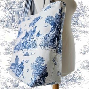 Può includere: Una borsa tote bianca con una stampa toile de Jouy blu raffigurante una scena pastorale con alberi, figure e uccelli. La borsa ha una tracolla a righe blu navy e bianche. La borsa è esposta su un manichino.