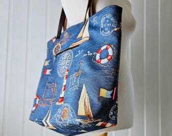 May include: A blue tote bag featuring a nautical print with sailboats, lighthouses, life preservers, compasses, and maritime signal flags. The bag has dark brown leather-like shoulder straps and a zippered exterior pocket on the front side.