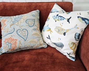May include: Two decorative throw pillows on a rust-colored couch. One pillow has a floral pattern in shades of blue, brown, and orange. The other pillow has a white background with a blue, yellow, and orange fish pattern.