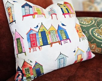 May include: A white throw pillow with a colorful pattern of beach houses. The houses are painted in various shades of blue, green, red, yellow, and pink. Each house has a unique design and features a small, white bird perched on the roof.