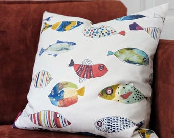 May include: A white square pillow with a colorful fish pattern. The fish are in various colors, including blue, red, yellow, and green. The pillow is resting on a dark red couch.