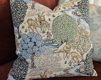 May include: A cream-colored throw pillow with a tapestry design featuring blue, green, and brown trees, a river, and deer. The pillow has a white border.