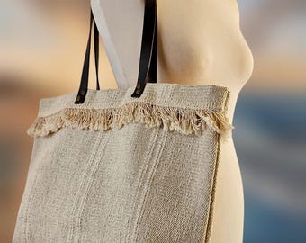 May include: A beige tote bag with black leather handles and a fringed top. The bag is displayed on a mannequin and has a woven texture with vertical stripes. The bag is designed for carrying items.